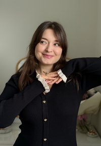 Jeune femme en cardigan côtelé noir aux poignets blancs, souriante et regardant sur le côté, mains jointes près du menton, fond neutre.