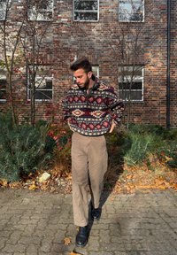 Strikket pullover i mørkerød og creme geometrisk mønster, kombineret med lysebrune bukser og sorte ankelstøvler, sat op mod en murstensbaggrund.