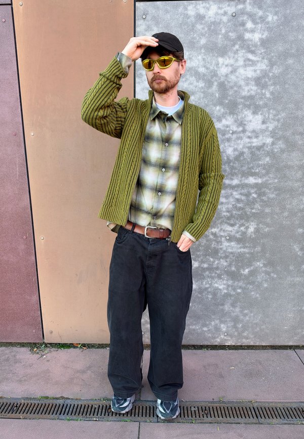 Un hombre con barba, gorra negra y gafas de sol amarillas se ajusta la gorra. Lleva una chaqueta de punto verde, una camisa de cuadros, vaqueros oscuros y zapatillas deportivas.