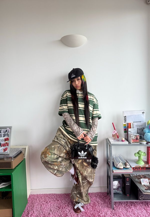 Person mit langen Haaren, die ein gestreiftes Shirt, Tarnhosen, eine Adidas-Kappe trägt und eine schwarze Adidas-Tasche hält, steht in einem Raum mit Regalen und Dekoration.