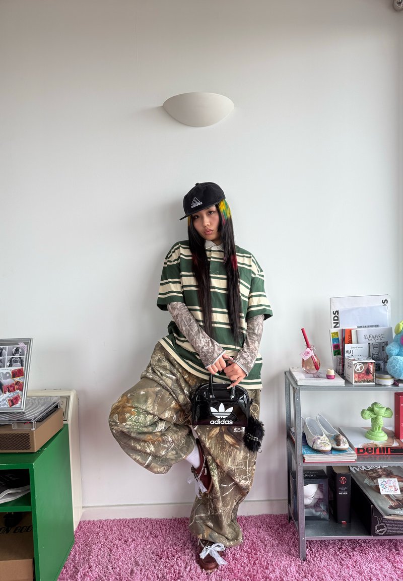 Personne aux cheveux longs portant une chemise rayée, un pantalon camouflage, une casquette Adidas et tenant un sac noir Adidas, debout dans une pièce avec des étagères et de la décoration.