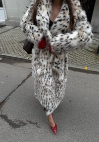 Viltotas kažokādas mētelis ar baltu un melnu plankumainu rakstu, pilna garuma, kombinēts ar sarkaniem cimdiem un sarkaniem augstpapēžu apaviem, stāvot pilsētas ielā.