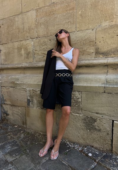 Blazer negro sobre una camiseta sin mangas blanca, combinado con shorts negros asegurados por un cinturón circular dorado; adornado con chanclas transparentes.