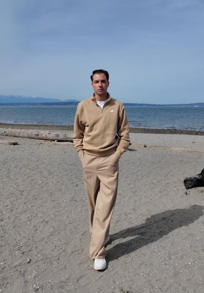 Joven con sudadera y pantalones beige de Nike caminando por una playa de arena con agua tranquila y montañas al fondo.