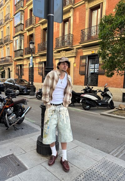 Hombre con gorra, camisa de cuadros, camiseta blanca y pantalones cortos largos decorados apoyado en una farola en una zona urbana.