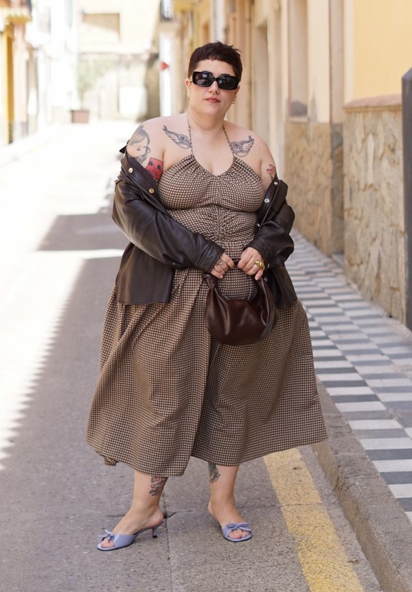Persoon die een zonnebril, een bruine geruite jurk, een zwarte leren jas en blauwe sandalen draagt, staat op straat en houdt een bruine handtas vast.