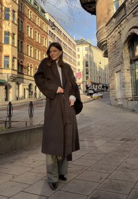 Long manteau marron serré à la taille, pull gris en dessous, et pantalon à rayures fines. Chaussures noires pointues, cadre urbain avec des bâtiments en arrière-plan.