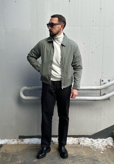 Chaqueta ligera gris, suéter de cuello alto blanco, pantalones negros y zapatos negros. La chaqueta tiene un cierre con cremallera y bolsillos laterales.