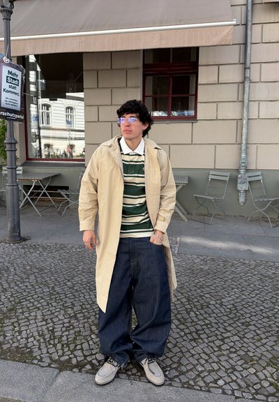 Joven con cabello rizado, gafas, abrigo beige, camisa a rayas, vaqueros anchos oscuros y zapatos grises, de pie en una calle empedrada.