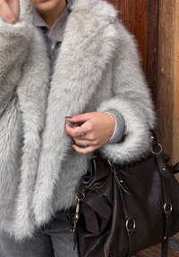 Szürke műszőrme kabát széles gallérral, szürke pulóver fölött viselve. Sötét bőrtáska rézszínű fém díszítéssel az alkaron pihen, lágy textúrát mutatva.