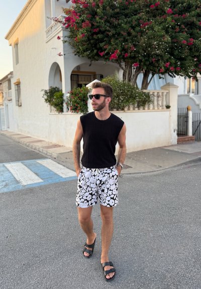 Top sin mangas negro, pantalones cortos negros y blancos con diseño floral, sandalias negras; de pie en un paso de peatones cerca de un edificio blanco.