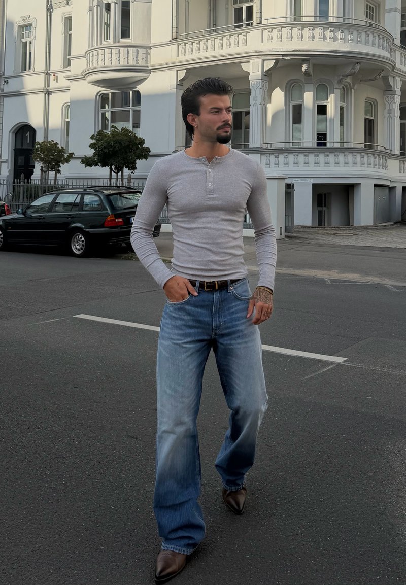 Uomo con capelli scuri e barba, indossa una maglia grigia a maniche lunghe, jeans blu e scarpe marroni, camminando in una strada della città vicino a un edificio bianco.