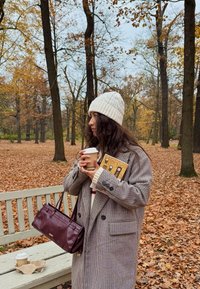 Manteau pied-de-poule surdimensionné, bonnet en tricot crème, sac à main bordeaux, tenant une tasse de café et un carnet, feuilles d'automne en arrière-plan.