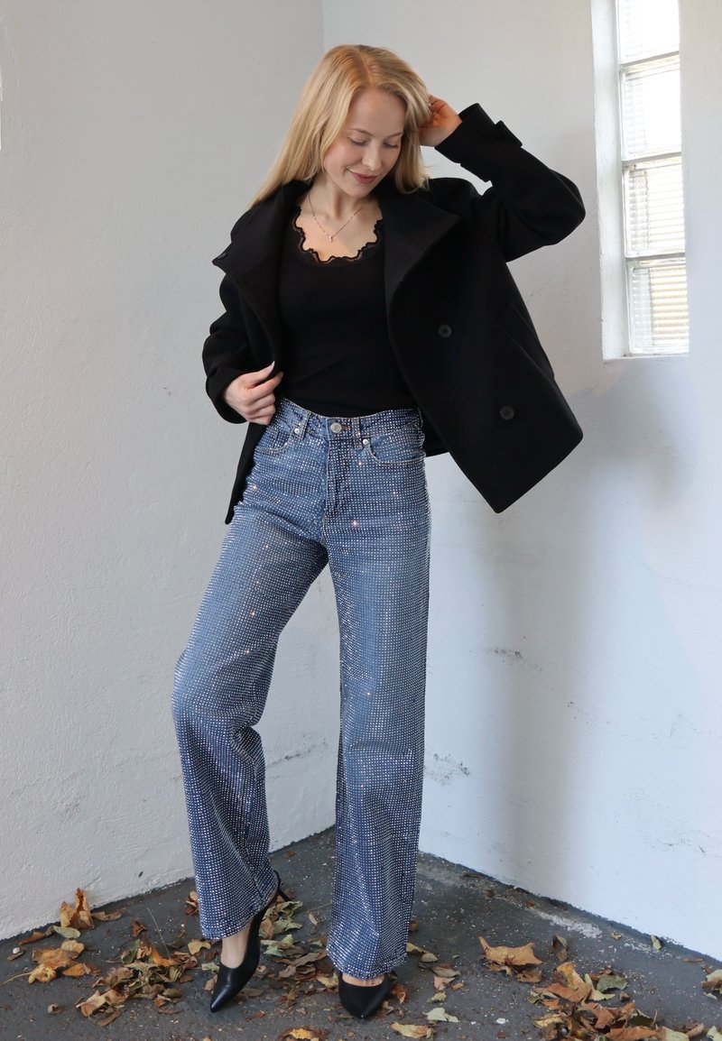 Black double-breasted coat over a fitted black top and sparkling blue wide-leg jeans; pointed black heels, textured fabric with embellishments.