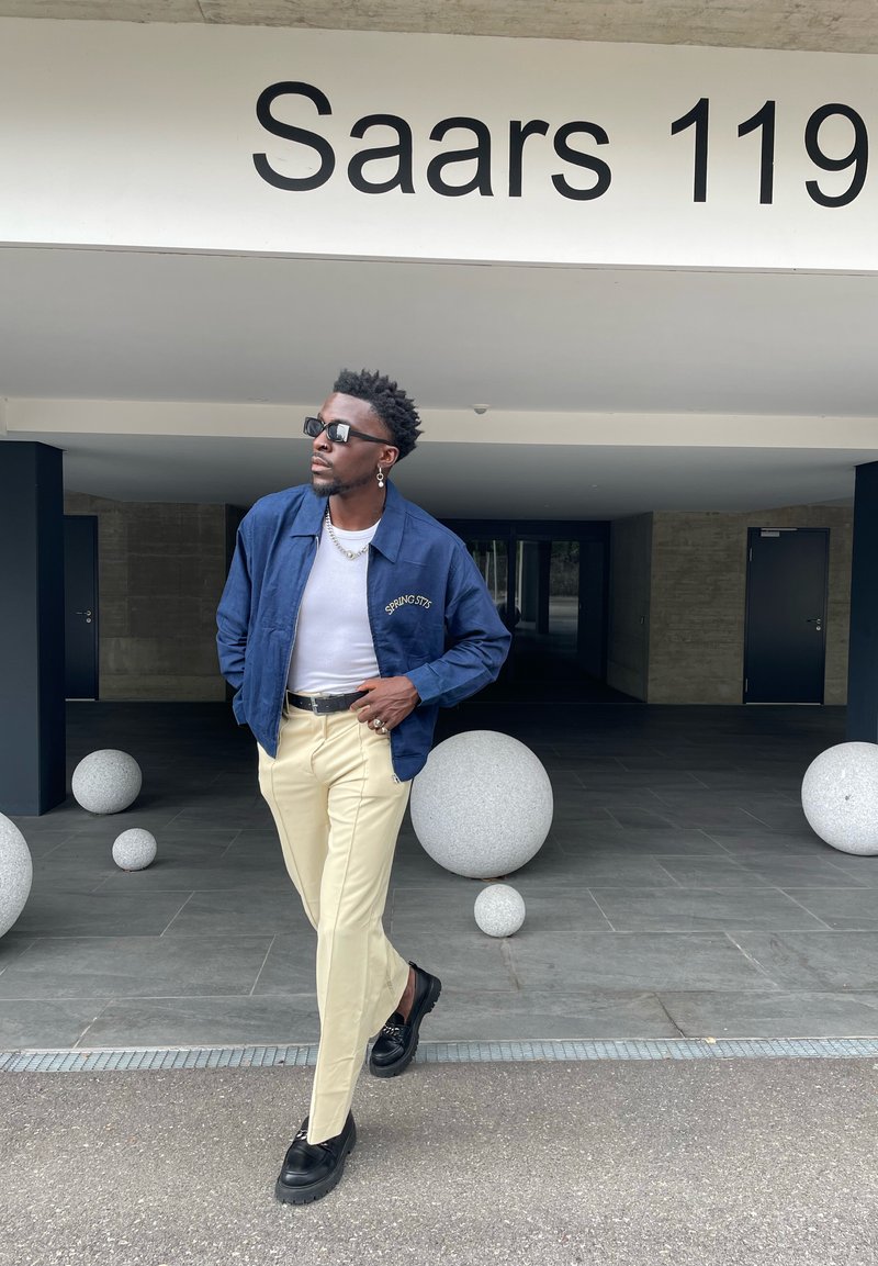 Homem com um casaco azul marinho, camisa branca, calças bege e sapatos pretos a caminhar perto de grandes esferas de pedra sob uma placa do edifício com a inscrição "Saars 119".