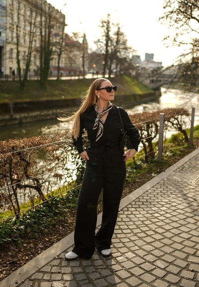 Conjunto de camisa negra y pantalones de pierna ancha con cintura ajustada. Los accesorios incluyen gafas de sol de gran tamaño y una bufanda estampada alrededor del cuello. Zapatillas blancas.