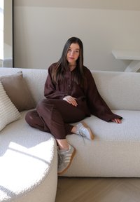 Καφέ λινό τοπ και παντελόνι με χαλαρή γραμμή, σε συνδυασμό με γκρι αθλητικά παπούτσια με λευκές ρίγες. Υφασμάτινος λευκός καναπές σε ανοιχτό ξύλινο δάπεδο.