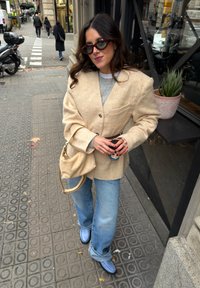 Femme en veste beige et jean bleu clair debout sur le trottoir de la ville, tenant une tasse de café, portant des lunettes de soleil et un sac beige assorti.