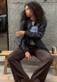 Kvinde med krøllet hår, der sidder på en bænke i træ mod en stenmur, iført en pinstribet blazer med stort blåt blomsteraccessorie og brune brede bukser.