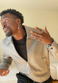 Muž s náušnicí a prsteny gestikuluje rukama, má na sobě šedou bundu na zip a černé tričko, stojí před béžovou zdí.