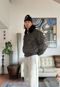 Blouson aviateur noir rembourré avec col en fourrure, pantalon de survêtement gris et bonnet noir. Debout dans un cadre intérieur moderne avec des éléments de décoration.