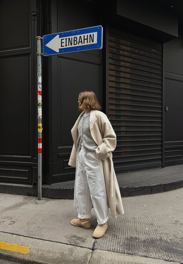 Personne portant un manteau beige, un pull gris, un pantalon blanc et des chaussures beiges, debout sur le trottoir sous un panneau de signalisation bleue indiquant une rue à sens unique « Einbahn ».