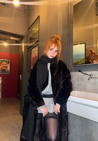 Dlhý čierny kabát z umelého kožušiny cez sivú mikinu, čierny opasok v páse, šortky s čipkovaným lemom a čierne pančuchy. Tmavý, moderný interiér.