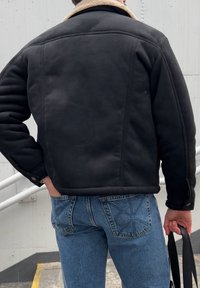 Veste noire avec col en shearling, texture matelassée, fermeture à boutons et poches latérales, associée à un jean en denim bleu clair.