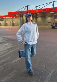 Sweat-shirt gris par-dessus une chemise blanche à col, jean bleu ample et casquette bleue. Le sujet est debout sur une jambe, avec une station-service en arrière-plan.