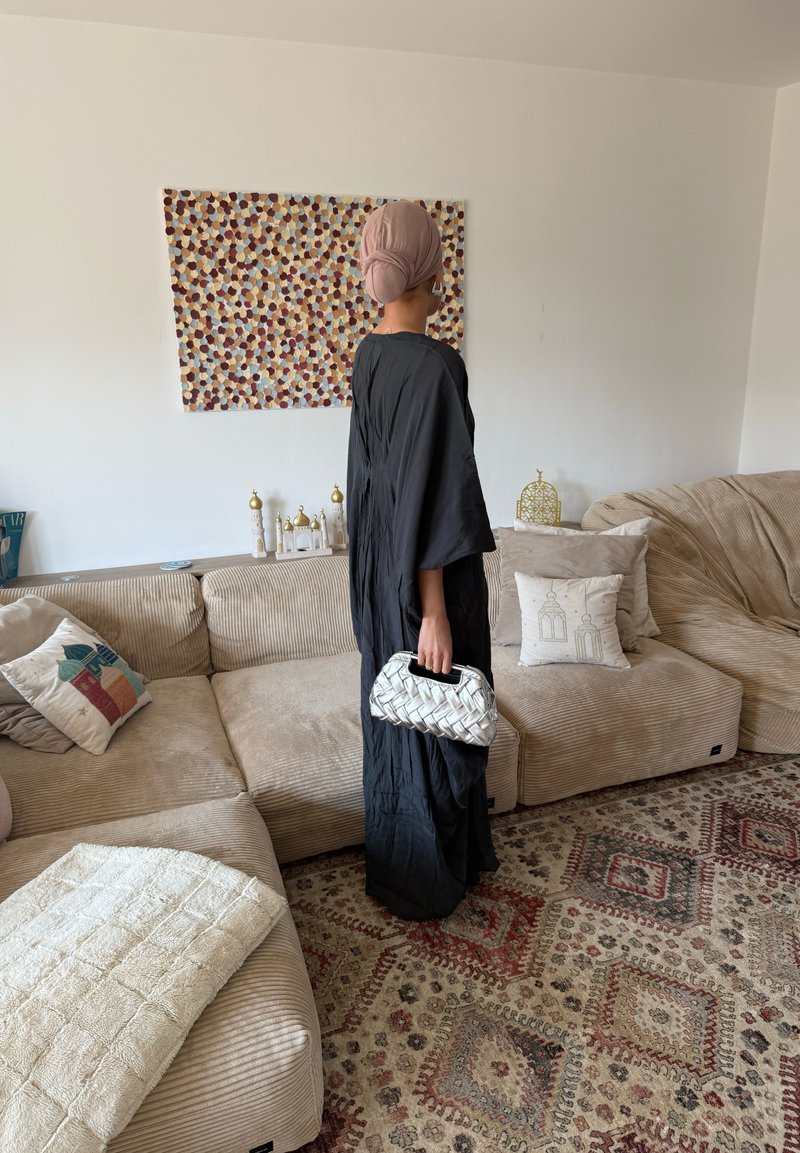 Eine Frau trägt eine schwarze Abaya und ein beigefarbenes Kopftuch, steht in einem Wohnzimmer und hält eine weiße gewebte Handtasche, während sie einem gemusterten Wandbehang gegenübersteht.