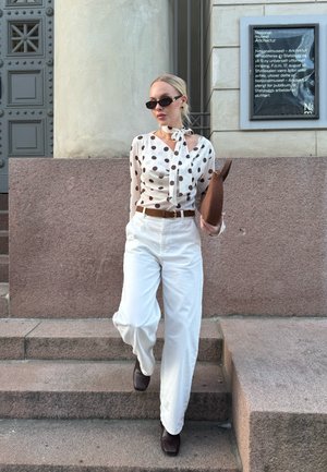 Mujer con gafas de sol, blusa de lunares, pantalones blancos de pierna ancha y botas marrones, sosteniendo un bolso de mano marrón mientras baja unas escaleras de piedra.