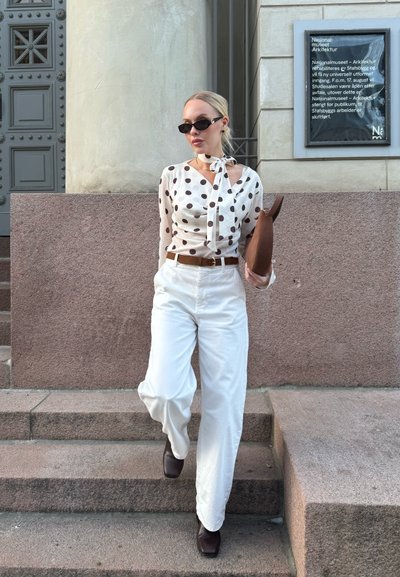 Mujer con gafas de sol, blusa de lunares, pantalones blancos de pierna ancha y botas marrones, sosteniendo un bolso de mano marrón mientras baja unas escaleras de piedra.