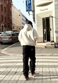 Person iført en hvid hættebluse og sorte bukser krydser et zebra-stribet vej i en by; bygninger og parkerede biler i baggrunden.