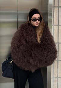 Manteau oversize marron duveteux avec un extérieur texturé et bouclé. Porté avec un bandeau noir et des lunettes de soleil, associé à un sac noir.