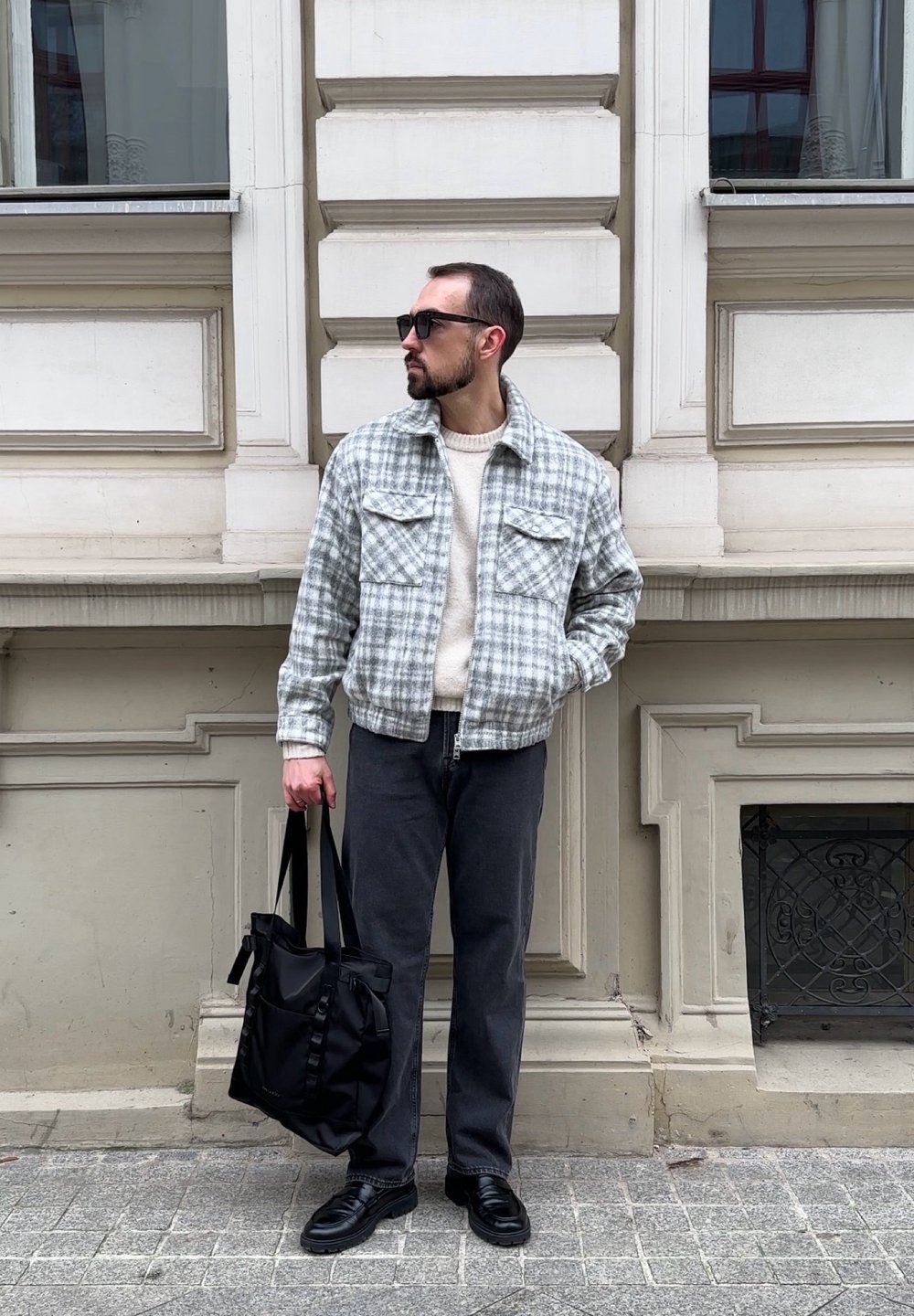 Giacca a quadri grigia cropped sopra un maglione beige, pantaloni grigio scuro e scarpe nere. Tiene una borsa tote nera, in piedi contro un muro texturizzato.