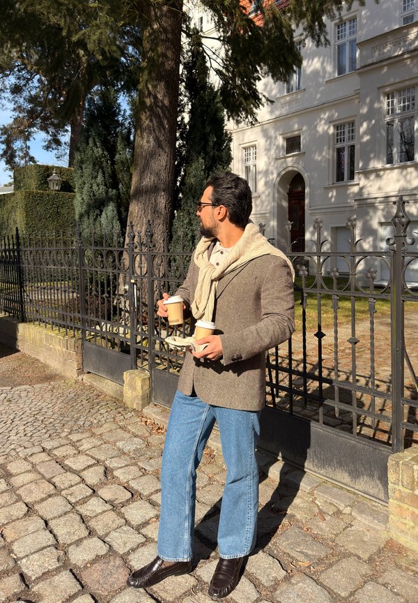 Mann med briller og skjerf som holder tre takeaway-kopper, står på brosteinsbelagt gate ved et svart smijernsstak nær en hvit bygning.