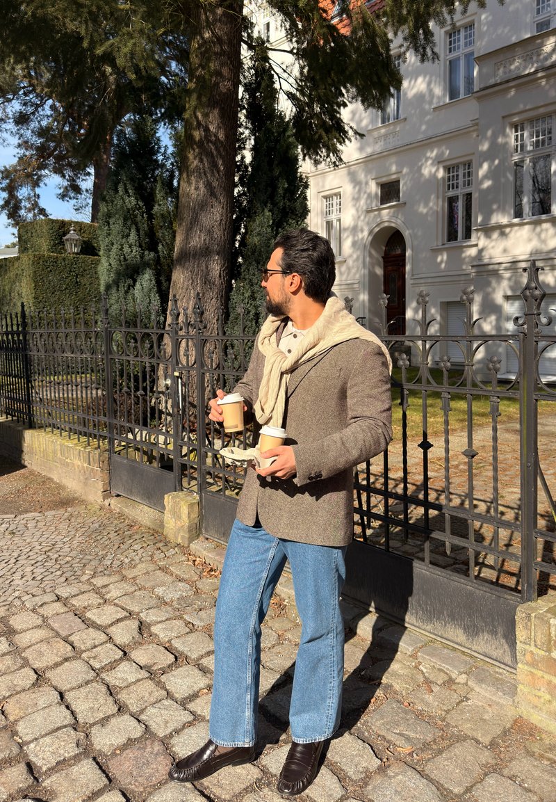 Άνδρας με γυαλιά και κασκόλ κρατά τρεις χάρτινες κούπες καφέ για πακέτο, στέκεται σε πλακόστρωτο δρόμο κοντά σε μαύρο σιδερένιο φράχτη δίπλα σε λευκό κτήριο.