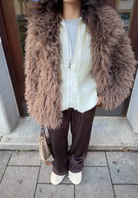 Veste extérieure marron et duveteuse portée sur un chemisier léger rayé ; associée à un pantalon sombre et fluide et des chaussures crème. Un sac à main à motif est tenu à la main.