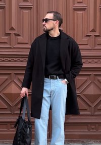 Černý kabát přes černý svetr, světle modré džíny a černá taška. Doplňky zahrnují sluneční brýle a hodinky. Pozadí tvoří hnědé dveře.