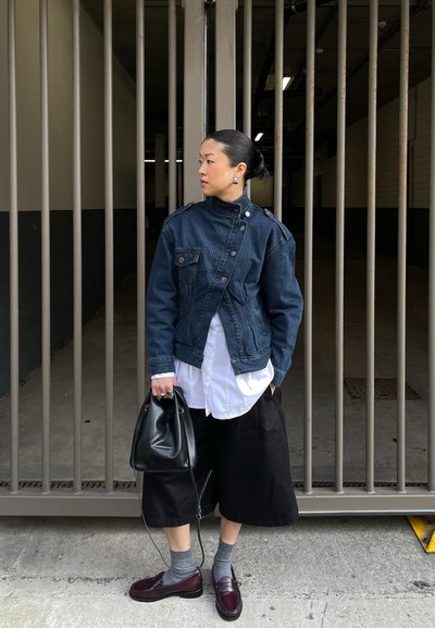 Mujer con cabello negro recogido en moño, vistiendo chaqueta de mezclilla oscura, camisa blanca, pantalones anchos negros, calcetines grises y zapatos burdeos, sosteniendo un bolso negro junto a una puerta metálica.