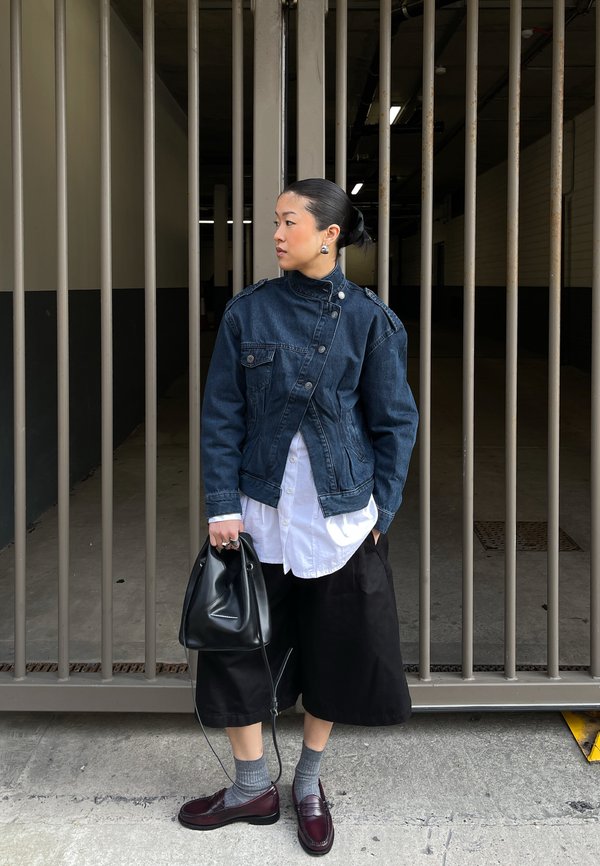 Nainen, jolla on mustat hiukset nutturalla, pukeutuneena tummansiniseen farkkutakkiin, valkoiseen paitaan, mustiin leveälahkeisiin housuihin, harmaisiin sukkiin ja viininpunaisiin kenkiin, pitää mustaa käsilaukkua metalliaidan vieressä.