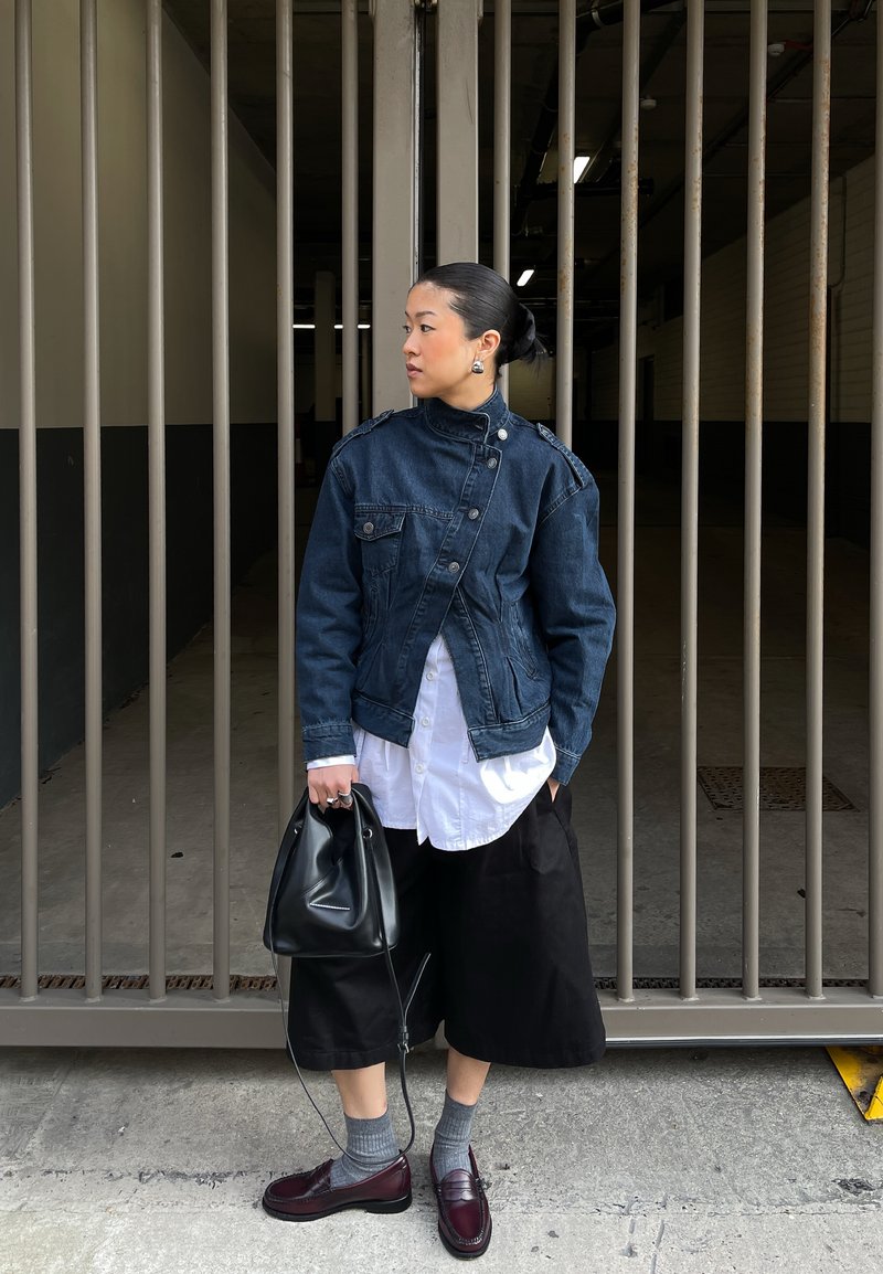 Mustade juustega naine kuklas, kandes tumedat teksajopet, valget särki, musti laiu pükse, halli sokke, burundukivärvi kingi, hoides metallvärava ääres musta käekotti.