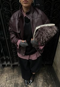 Veste en cuir bordeaux portée sur un pull noir, associée à une chemise à rayures roses, un pantalon noir à jambes larges et des gants gris en peluche. Tenant un livre.
