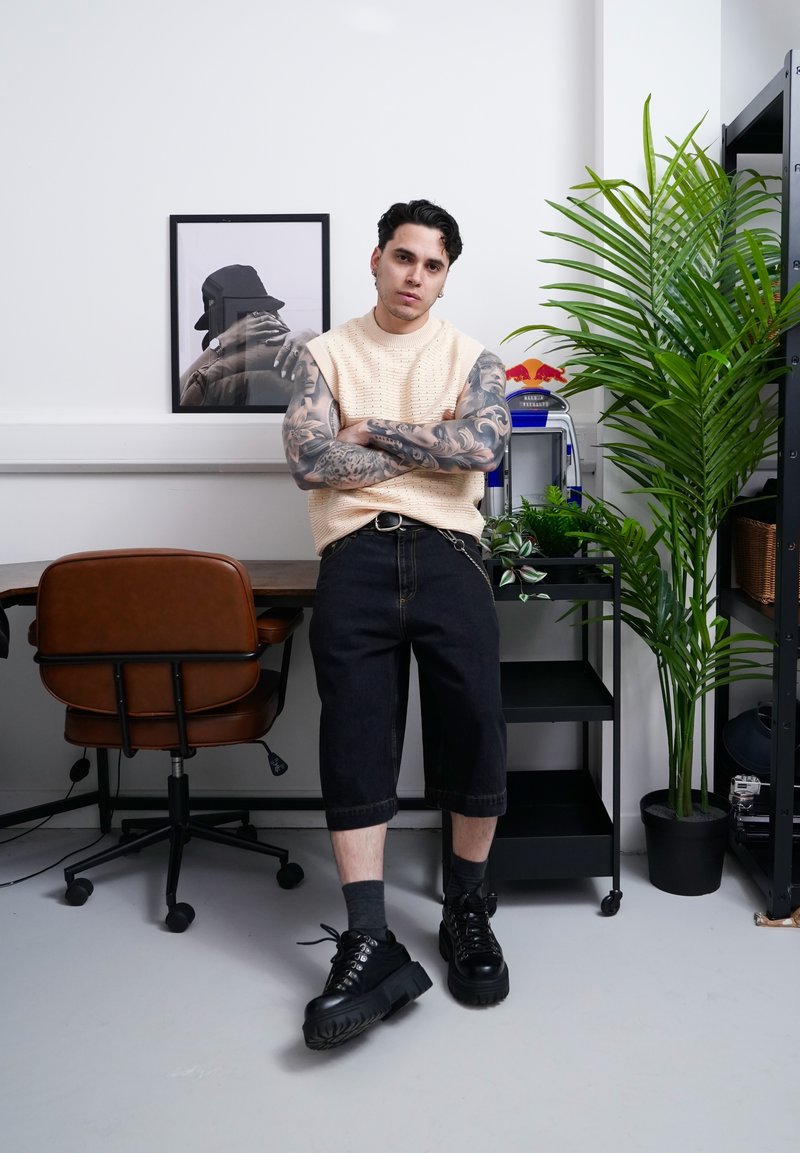 Man met getatoeëerde armen, mouwloze beige trui en zwarte short leunend tegen een bureau in een modern vertrek met planten en ingelijste foto.