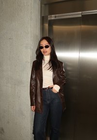 Γυναίκα με μακριά σκούρα μαλλιά, φορώντας γυαλιά ηλίου, λευκό ζιβάγκο, καφέ δερμάτινο μπουφάν και σκούρο τζιν, στέκεται δίπλα σε κλειστή πόρτα ανελκυστήρα.