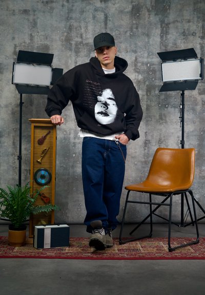 Sudadera negra con capucha y estampado gráfico de un rostro, combinada con jeans azules holgados y botas grises. Cerca hay una silla marrón y objetos decorativos.
