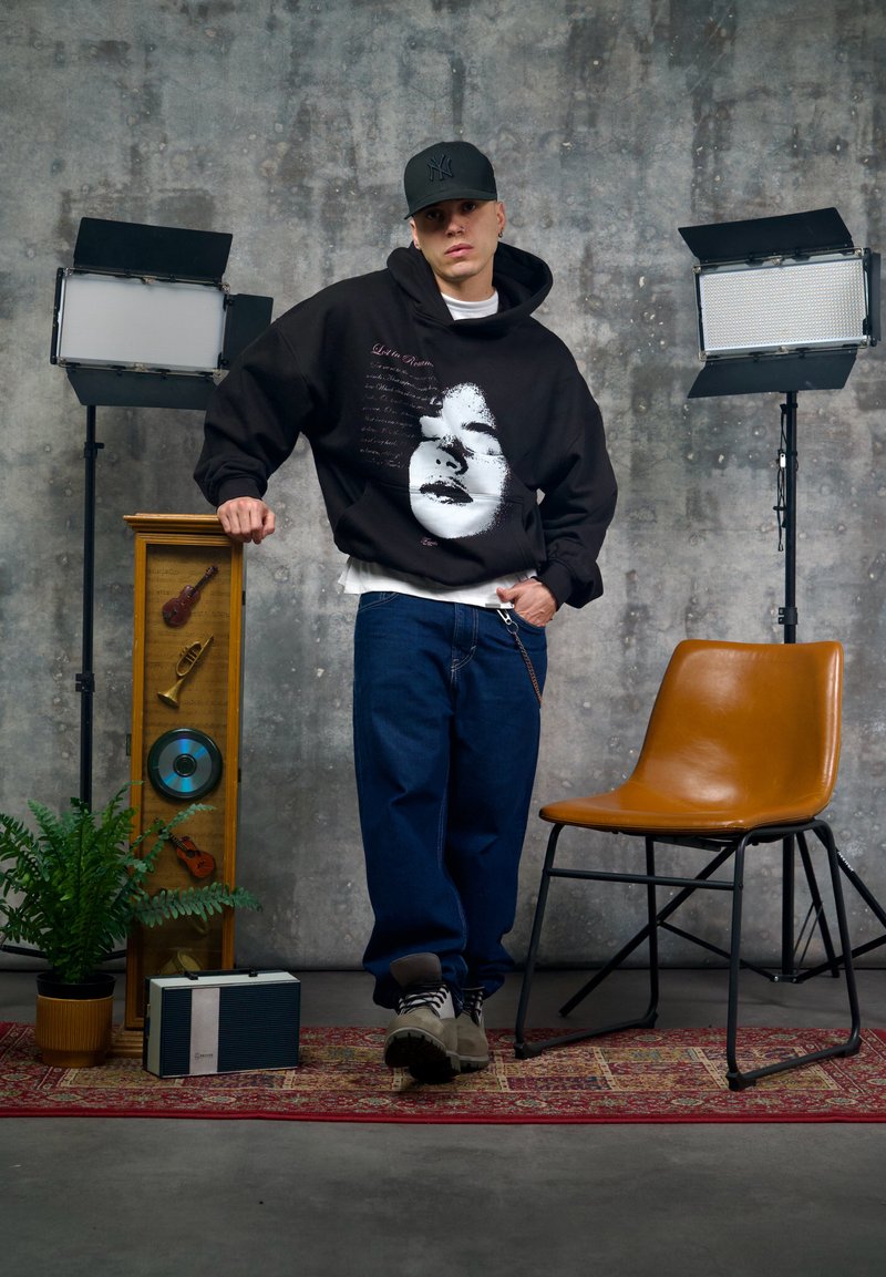 Schwarzer Kapuzenpullover mit einem grafischen Gesichtsdruck, kombiniert mit lockeren blauen Jeans und grauen Stiefeln. Ein brauner Stuhl und dekorative Gegenstände befinden sich in der Nähe.