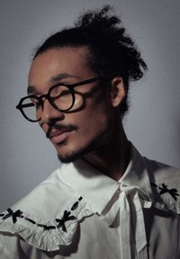 Lunettes à monture noire, blouse blanche avec col à volants, accents noirs et une finition lisse et texturée ; cheveux bouclés attachés lâchement.