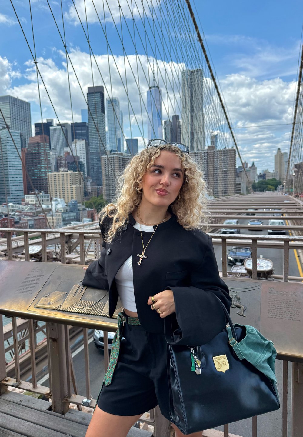 Svart oversized jakke, hvit crop-top og svarte shorts. Stor, to-farget håndveske med lapper og nøkkelringer. Urbant miljø med skyline i bakgrunnen.
