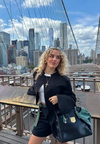 Svart oversized jakke, hvit crop-top og svarte shorts. Stor, to-farget håndveske med lapper og nøkkelringer. Urbant miljø med skyline i bakgrunnen.